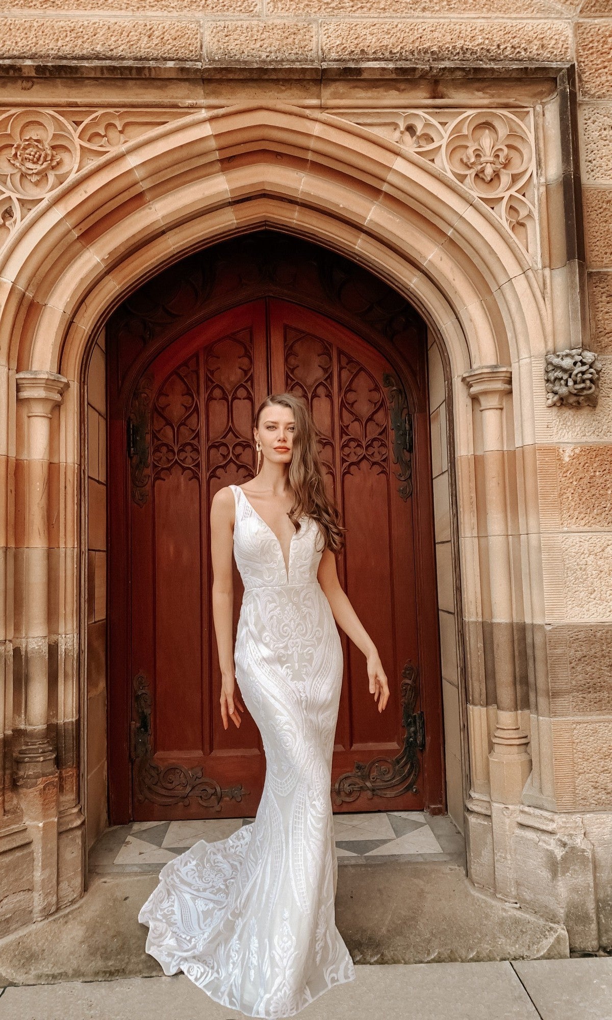 A woman in the Tina Holly BA109 White Lace Mermaid Wedding Gown stands before an ornate arched wooden door set in a stone building.